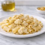 In primo piano su un tavolo di marmo chiaro c'è un piatto di pasta con cavolfiore con del formaggio e noce moscata. Sulla destra c'è un tovagliolo grigio con una forchetta.