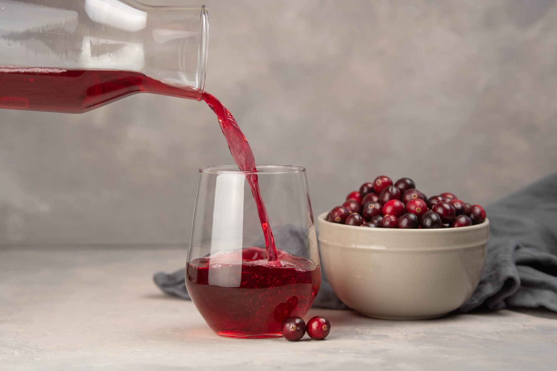 mano fuoricampo che versa da una brocca un succo di mirtillo rosso i bicchiere. Dietro ciotola piena di bacche di mirtillo rosso