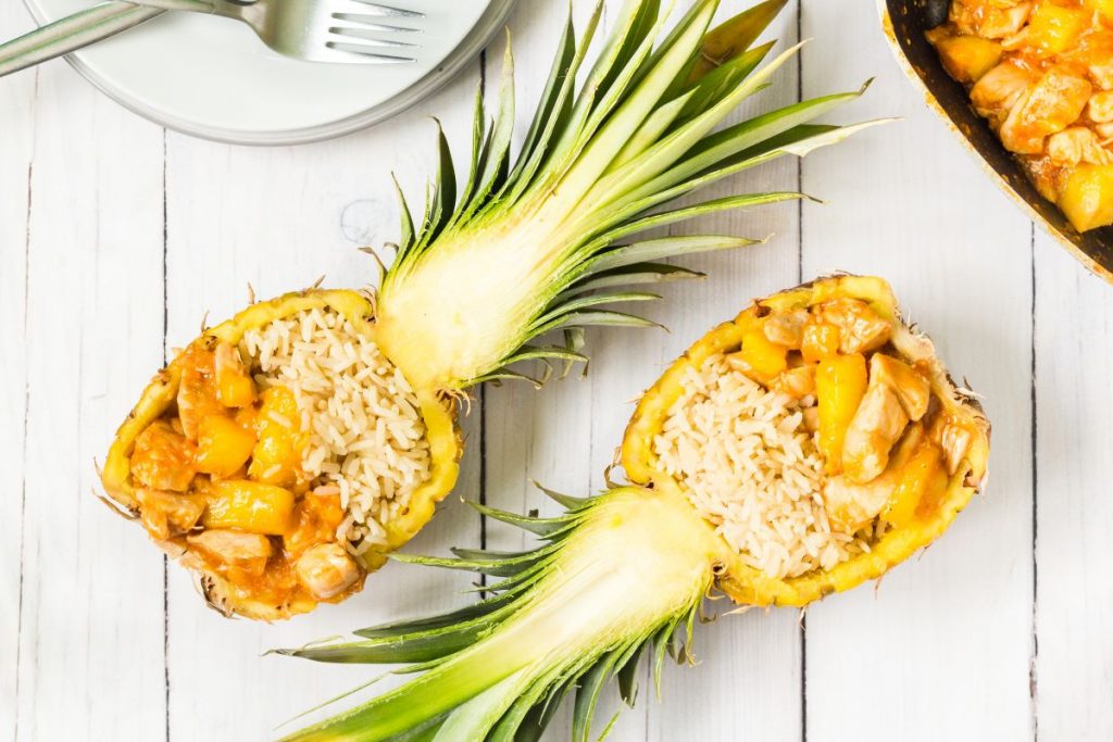 Pollo all'ananas, un classico amatissimo della cucina orientale 2 su base di legno chiaro due metà di un ananas ripiene di riso e bocconcini di pollo