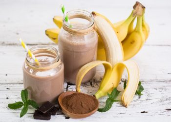 su sfondo bianco 2 bicchieri con frullati di banana e banane sbucciate e cacao amaro