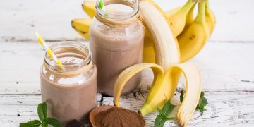 su sfondo bianco 2 bicchieri con frullati di banana e banane sbucciate e cacao amaro