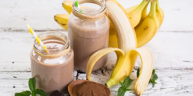 su sfondo bianco 2 bicchieri con frullati di banana e banane sbucciate e cacao amaro