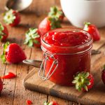 Marmellata di fragole, tre ricette facili per una dolce colazione 1 In primo piano su un tagliere di legno c'รจ un barattolo in vetro con della marmellata di fragole. Sullo sfondo delle fragole fresche e un cucchiaino.