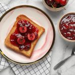 Marmellata di ciliegie, la confettura più deliziosa da fare in casa 1 In primo piano su un tavolo in marmo c'è un piatto con una fetta biscottata e della marmellata di ciliegie. Sulla destra una ciotolina con della marmellata e un cucchiaino.