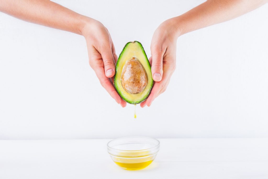 due mani femminili in primo piano che tengono in mano mezzo avocado e sotto una ciotolina con dell'olio di avocado
