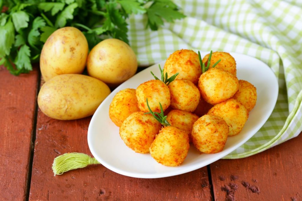 Polpette di patate: la ricetta con pochi ingredienti da provare subito! 1 su tavolo di legno, piatto bianco con polpette di patate, accanto 3 patate e dietro prezzemolo e canovaccio verde a quadretti bianchi