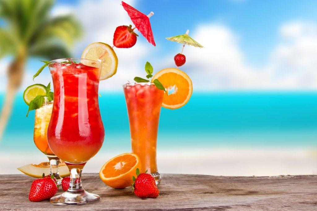 sulla sabbia con mare sul fondo 3 bicchieri colorati con cocktail, fette di frutta e bandierine