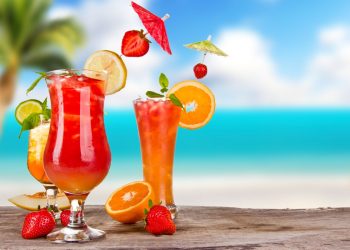 sulla sabbia con mare sul fondo 3 bicchieri colorati con cocktail, fette di frutta e bandierine