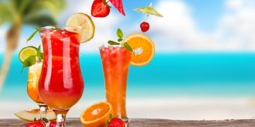 sulla sabbia con mare sul fondo 3 bicchieri colorati con cocktail, fette di frutta e bandierine