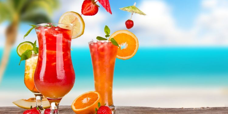sulla sabbia con mare sul fondo 3 bicchieri colorati con cocktail, fette di frutta e bandierine