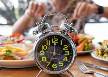 Donna che mangia su tavolo pieno di cibi con in primo piano un orologio per illustrare il digiuno intermittente