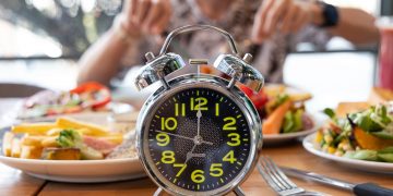 Donna che mangia su tavolo pieno di cibi con in primo piano un orologio per illustrare il digiuno intermittente