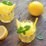 Granita al limone: la ricetta della granita siciliana fatta in casa 1 In primo piano due bicchieri di granita al limone con foglie di menta e spicchi di limone su un tavolo di legno, accanto a limoni freschi.