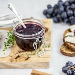 In primo piano sopra una tavoletta di legno, c'è un barattolo di vetro con la marmellata di uva nera, fette di pane con formaggio e chicchi d'uva.