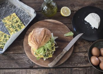 piatto scuro con un panino farcito di frittata e insalata, un coltello, uova e tranci di frittata