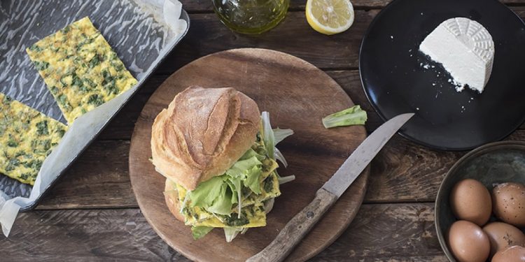 piatto scuro con un panino farcito di frittata e insalata, un coltello, uova e tranci di frittata