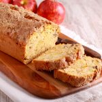 Plumcake alle mele e yogurt: la ricetta soffice senza burro 1 In primo piano su un tagliere di legno c'è un plumcake alle mele. Sullo sfondo delle mele rosse.
