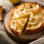 Torta di mele: ricetta facile con cannella e limone 1 In primo piano su un tagliere in legno c'รจ una soffice torta di mele. Sullo sfondo delle mele e delle stecche di cannella.