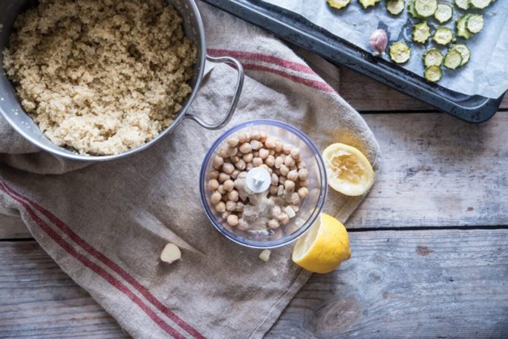In primo piano all'interno di un frullatore dei ceci per preparare l'hummus come salsa della Buddha bowl, accanto dei spicchi di limone e la pentola con della quinoa.