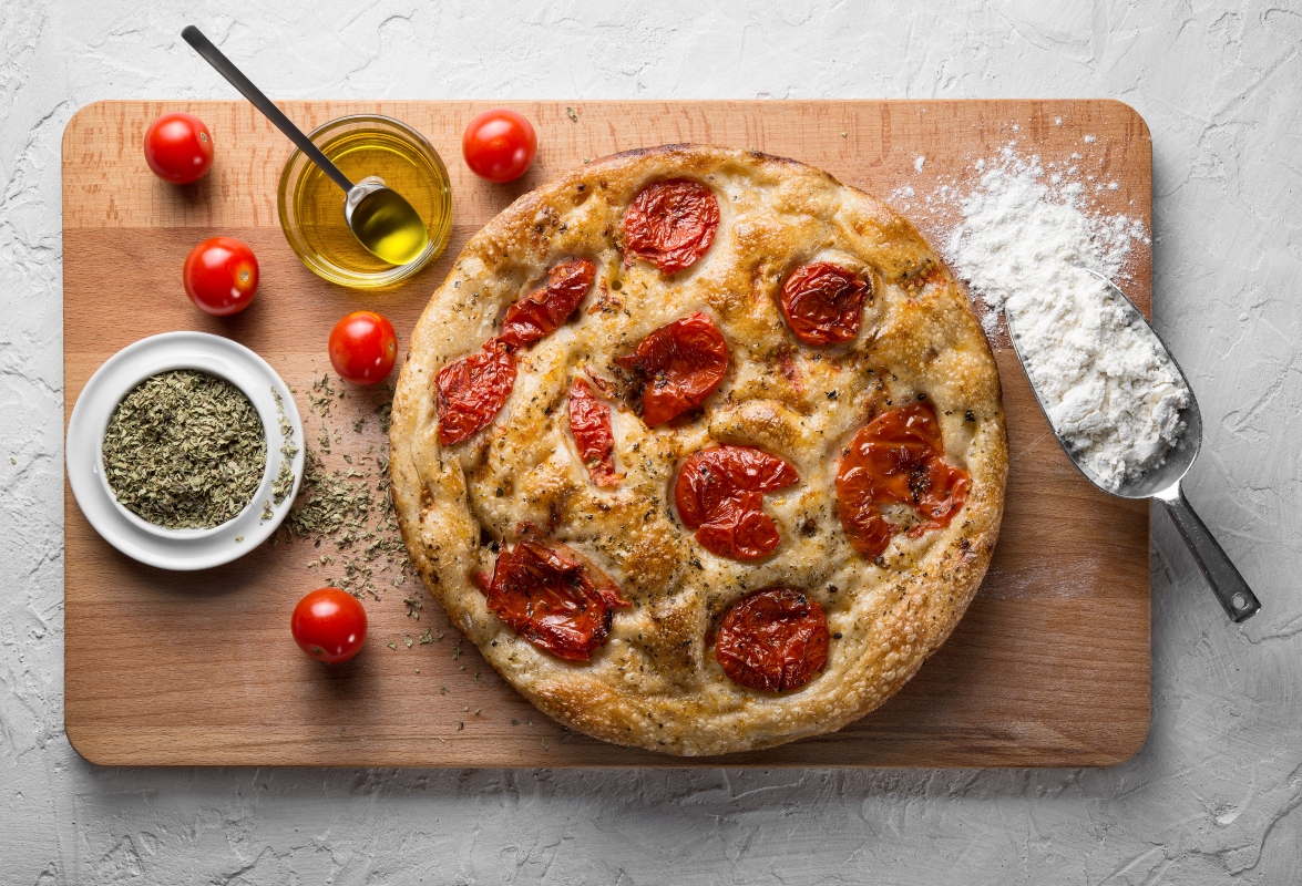 primo piano di un tagliere di legno su tavolo con una focaccia con pomodori con accanto nelle ciotole ingredienti come farina, pomodorini, una ciotola con olio EVO e erbe aromatiche