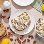 Insalata di pollo light: ricetta facile e veloce 1 su tavolo da cucina, contenitore con insalata di pollo light. Nel piatto accanto, piatto con pane integrale con l'insalata di pollo spalmata. Attorno, pane integrale, limoni e frutta secca