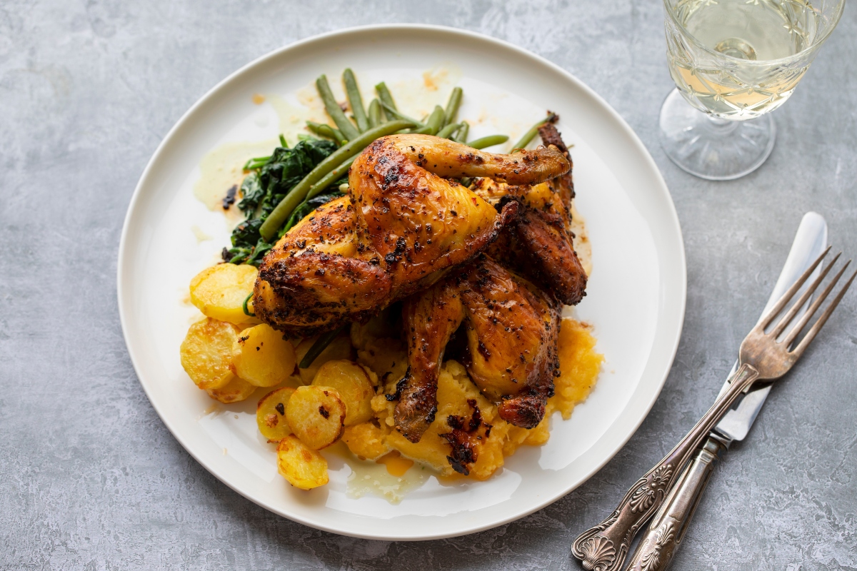 su tavolo, in cucina un piatto bianco visto dall'alto con pollo al forno con patate e fagiolini. Accanto bicchiere con vino bianco 1 forchetta e un coltello.