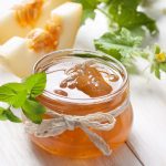 Marmellata di melone senza zucchero fatta in casa 1 In primo piano su un tavolo di legno bianco c'รจ un barattolo in vetro con della marmellata di melone. Sullo sfondo ci sono due fette di melone bianco.