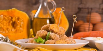 piatto bianco con polpette di zucca e spiedini, zucca aperta e flacone di olio sullo sfondo