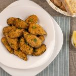Cozze gratinate al forno con panatura croccante 1 In primo piano su un tavolo di legno e una tovagliavette verde salvia a righe c'รจ un piatto fondo bianco con delle cozze gratinate. Sulla destra uno spicchio di limone e un cestino di pane.