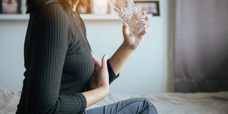 Donna seduta sul letto appoggia una mano sullo sterno perché ha dolore e nell'altra mano beve acqua nella bottiglia perché segue una dieta per reflusso