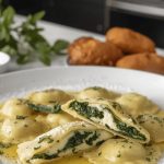 Ravioli ricotta e spinaci fatti in casa con ripieno cremoso e condimenti 1 In primo piano in un piatto fondo bianco dei ravioli ricotta e spinaci cremosi conditi con burro e salvia. Sullo sfondo ci sono alcuni ingredienti principali della ricetta come il parmigiano e la salvia.