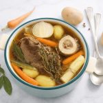 In primo piano su un tavolo di marmo chiaro c'è una pentola in ceramica bianca con del brodo di carne. Intorno c'è una foglia di allora, una carota, una patata e delle posate.