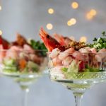 Cocktail di gamberi: l'antipasto perfetto per le tue feste con videoricetta 1 In primo piano due coppe di cristallo con cocktail di gamberi, alla base insalata, avocado e una fetta di lime. Sullo sfondo delle luci natalizie.