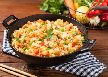 su tavolo di legno, un canovaccio a righe bianche e blu con baghette cinesi e un padella piena di riso alla cantonese, sulla destra cesto con verdure di stagione.
