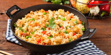 su tavolo di legno, un canovaccio a righe bianche e blu con baghette cinesi e un padella piena di riso alla cantonese, sulla destra cesto con verdure di stagione.