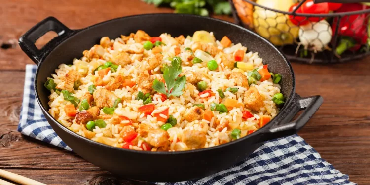 su tavolo di legno, un canovaccio a righe bianche e blu con baghette cinesi e un padella piena di riso alla cantonese, sulla destra cesto con verdure di stagione.