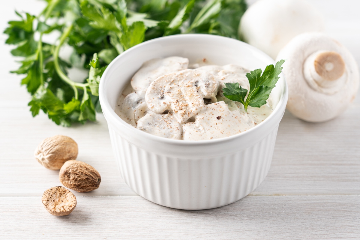 su tavolo bianco, primo piano di una ciotola con funghi tagliati e noce moscata grattugiata. A sinistra noce moscata intera, dietro prezzemolo e a destra un fungo
