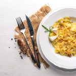 Pasta ai quattro formaggi: la ricetta per farla cremosa e avvolgente 1 In primo piano su un tavolo chiaro c'รจ un piatto fondo con la pasta ai quattro formaggi. Sulla sinistra delle posate su una tovaglietta in juta e dei grani di pepe nero.