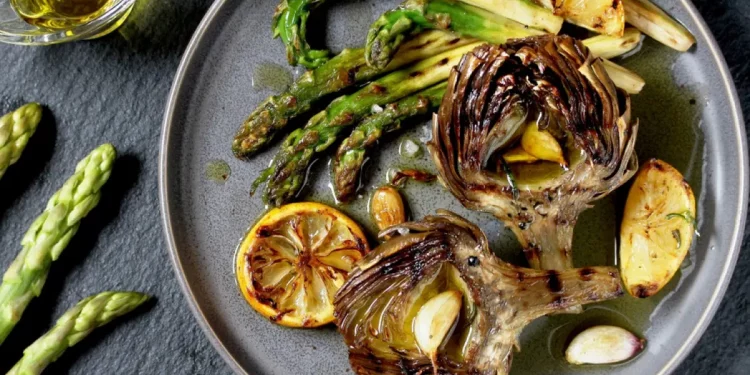 ricette con carciofi: su base scura un piatto tondo grigio con carciofi, asparagi e fette di limone