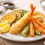 In primo piano su un piatto fondo chiaro c'è una tempura di verdure come zucchine, carote e cipolle e un gambero al centro. Sulla sinistra ci sono delle bacchette.