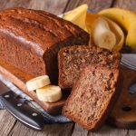 Banana bread senza glutine e senza lattosio: ricetta facile 1 su tavolo di legno, un canovaccio blu a righe bianche con il tagliere sopra e un banana bread senza glutine con 2 fette tagliate. Accanto 2 fette di banane e dietro una banana intera.