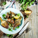 su tavolo di legno, piatto azzurro con 3 carciofi alla romana, forchetta e coltello accanto, mezzo lime e dietro sale e erbe aromatiche