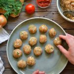 Polpette di pollo: ricetta leggera e morbida 1 su tavolo di legno da cucina piatto verde con polpette di pollo. Si vede la mano di una donna che le sistema nel piatto e accanto tutti gli ingredienti: cipolla, prezzemolo, pomodori e in un contenitore il macinato di pollo