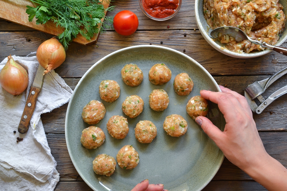 su tavolo di legno da cucina piatto verde con polpette di pollo. Si vede la mano di una donna che le sistema nel piatto e accanto tutti gli ingredienti: cipolla, prezzemolo, pomodori e in un contenitore il macinato di pollo