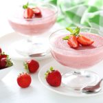 Mousse di fragole: la ricetta del dessert fresco e cremoso 1 In primo piano su un tavolo bianco c'è una coppa di vetro con della mousse di fragola e delle fragole fresche per decorare. Sullo sfondo un canovaccio a quadri verdi e bianchi.