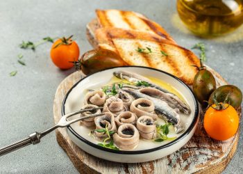 su tavolo, tagliere di legno con un piatto con pesce azzurro e attorno, 2 bruschette e pomodori arancioni