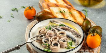 su tavolo, tagliere di legno con un piatto con pesce azzurro e attorno, 2 bruschette e pomodori arancioni