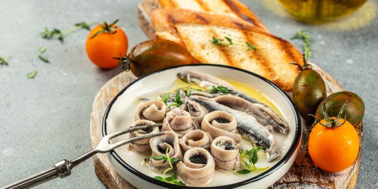 su tavolo, tagliere di legno con un piatto con pesce azzurro e attorno, 2 bruschette e pomodori arancioni