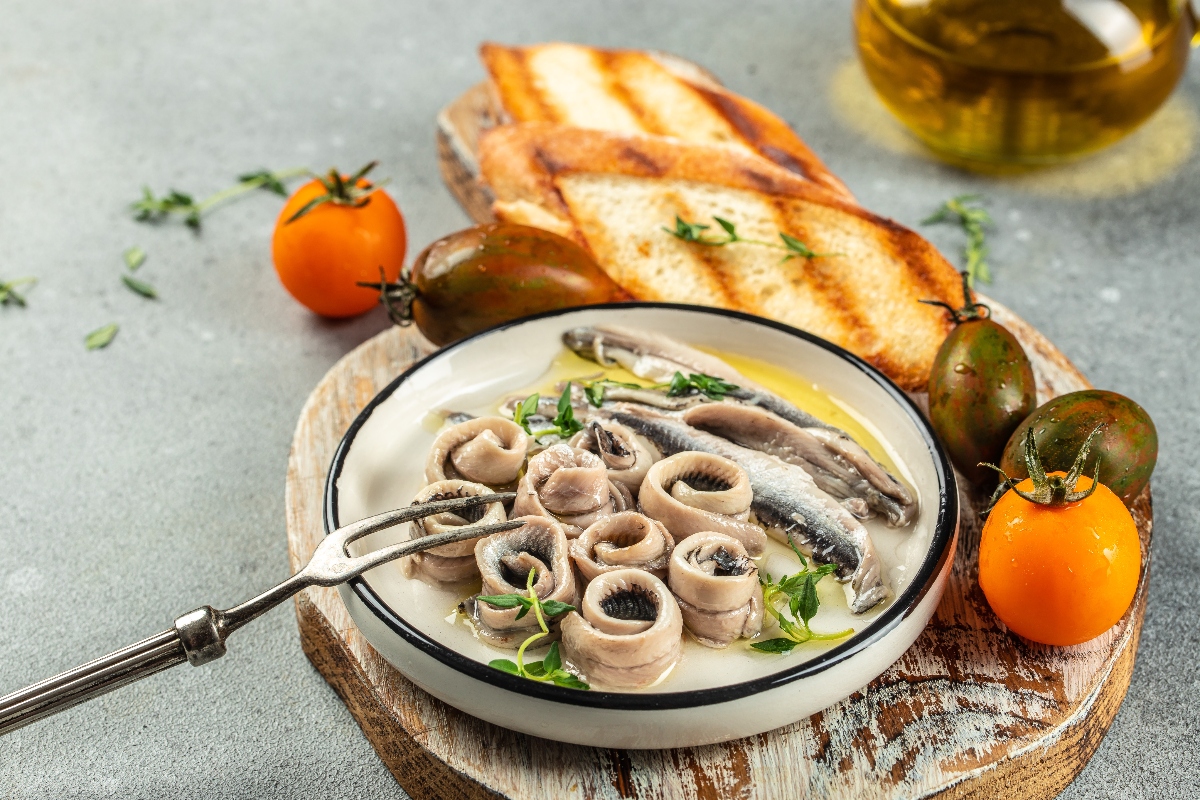 su tavolo, tagliere di legno con un piatto con pesce azzurro e attorno, 2 bruschette e pomodori arancioni