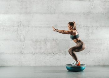 In palestra, con muro bianco, una donna con leggings e top con la coda di cavallo su la bosu ball esegue uno squat correttamente